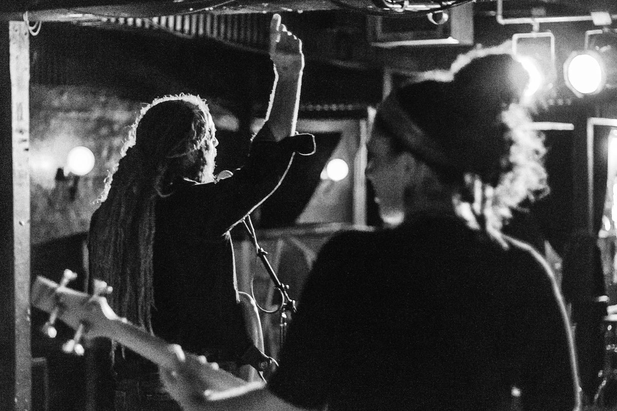 Black-and-white photo from behind a dreadlocked guitarist and a fan with a raised hand at a bar show.
