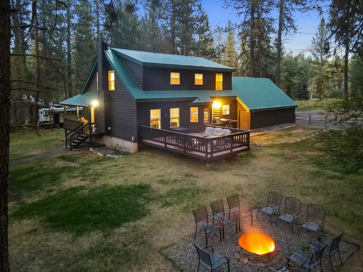 Drone view of a dark-sided cabin with lit windows at dusk, a fire pit glowing in the pine-lined yard.