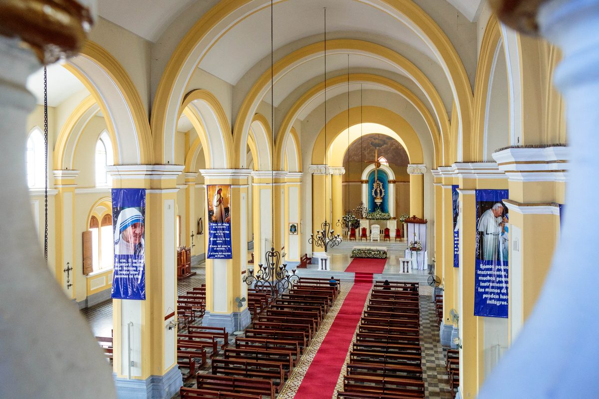 View from a balcony down a yellow-arched church nave with a red-carpeted aisle and saint banners.