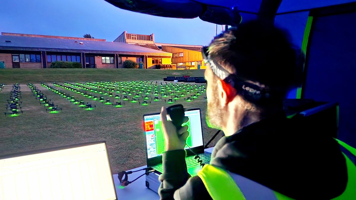 A drone show pilot in hi-vis at the control laptop watching a glowing green drone grid
