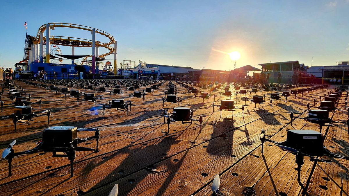 Rows of show drones set out on the sand with a pier and Ferris wheel at sunset