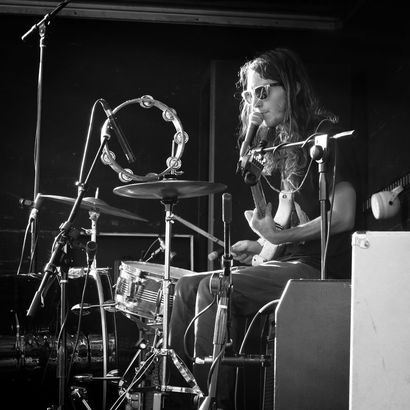 Black-and-white photo of a drummer playing a full kit with a tambourine in a small club