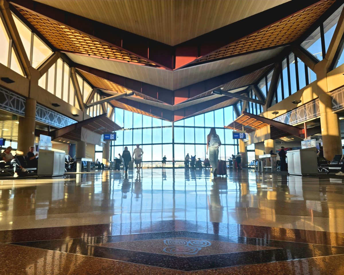 A bright glass airport terminal with a timber ceiling reflected in a polished floor