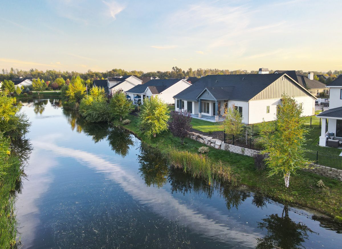 Large white and gray homes line a calm pond that mirrors the sky and trees.