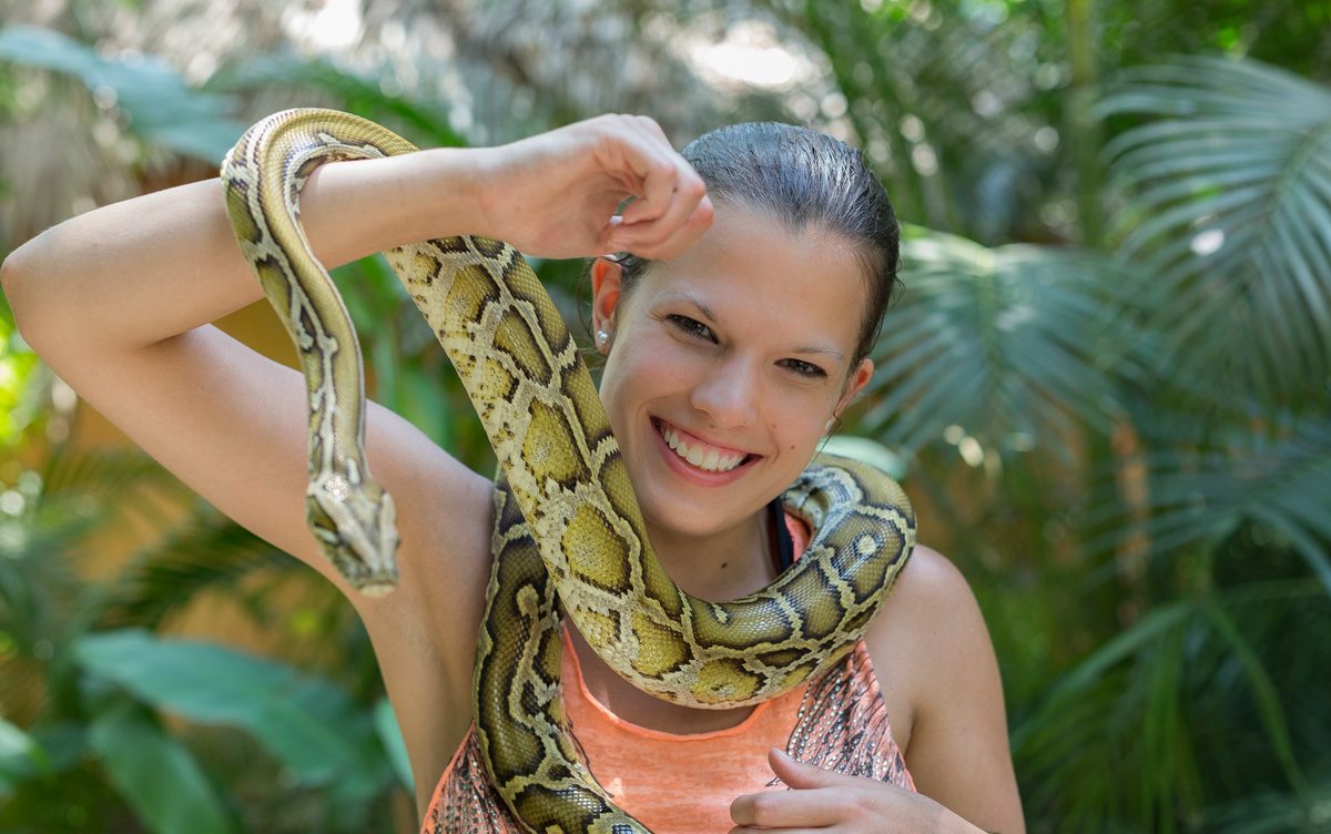 A smiling woman holding a large python draped around her shoulders in a tropical garden