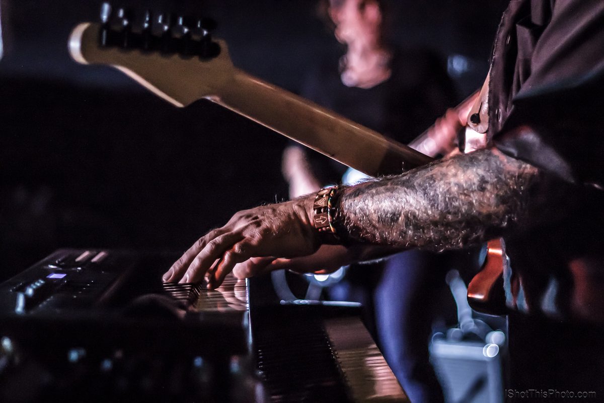 Close-up of a tattooed hand on a keyboard with a guitar neck behind, live at the Crazy Horse in Boise