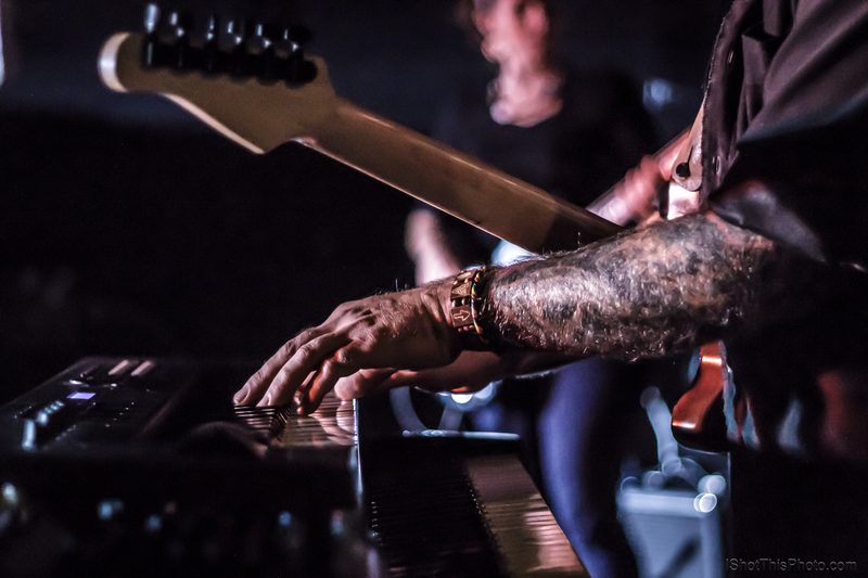Close-up of a tattooed hand on a keyboard with a guitar neck behind, live at the Crazy Horse in Boise
