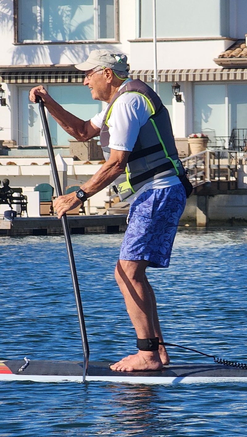 An older man in a life vest paddleboarding past harbor homes