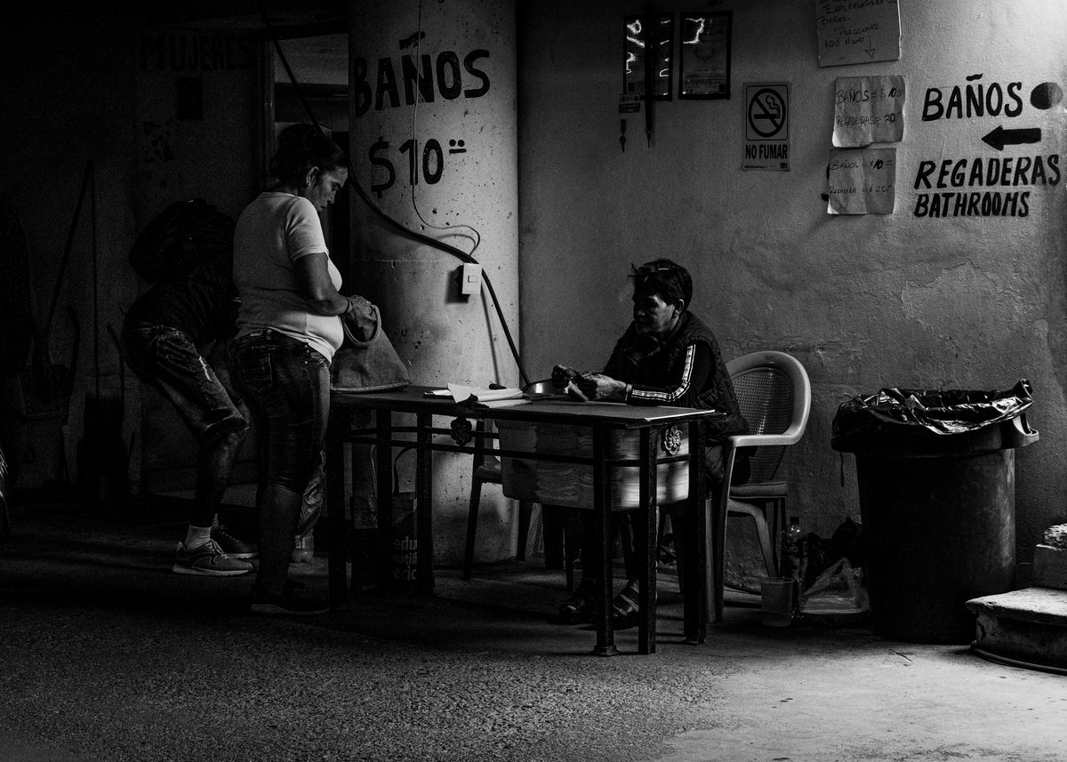 Black-and-white photo of a worn room with hand-painted baños signs and two people at a table.