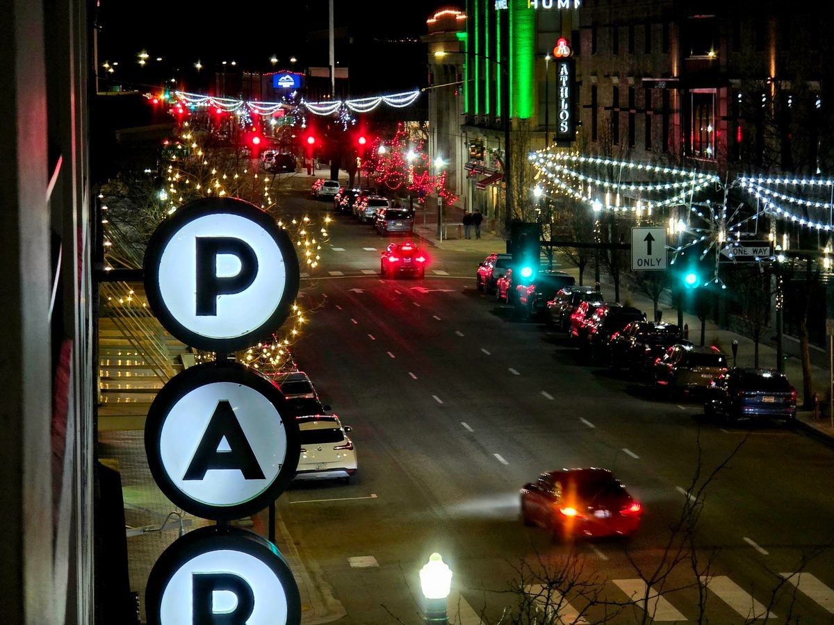 A glowing parking sign above a downtown street at night with string lights and red taillights