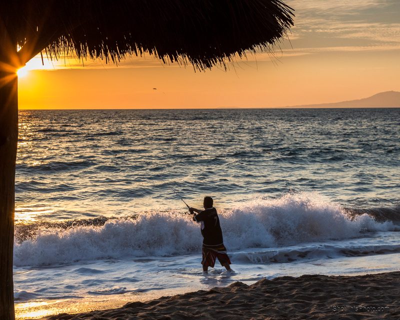 A man standing in breaking waves under a palapa roof at sunset in Puerto Vallarta