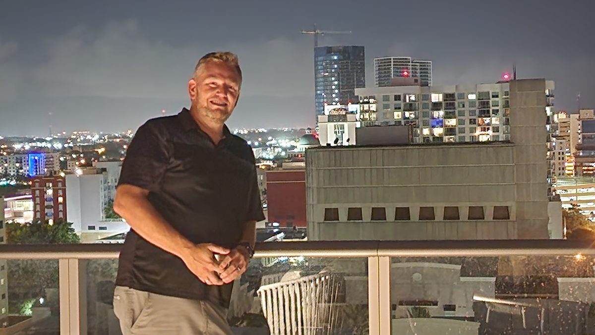 A man in a polo standing at a rooftop railing above city lights at night