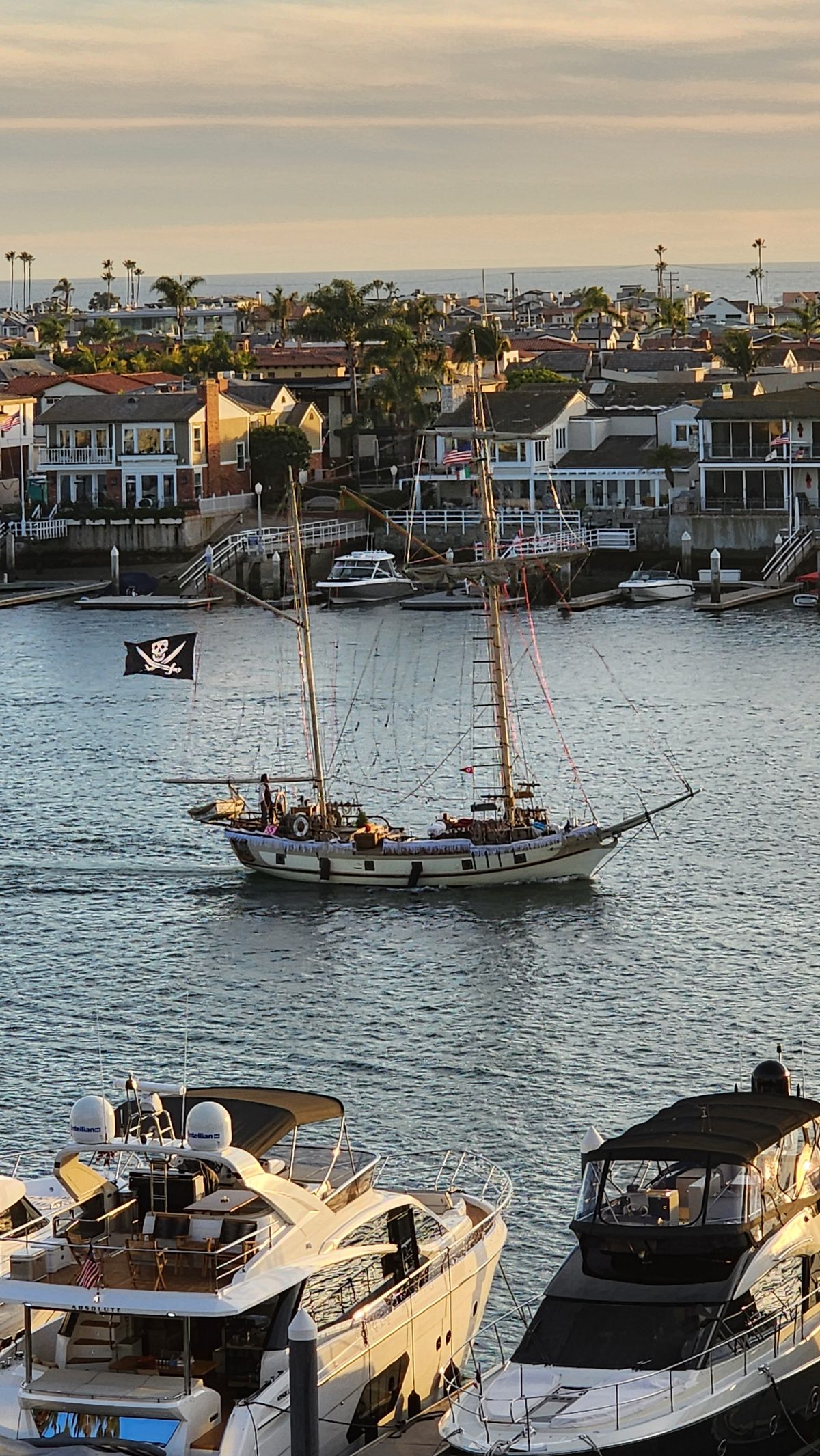 A two-masted schooner sailing past yachts in a harbor at golden hour