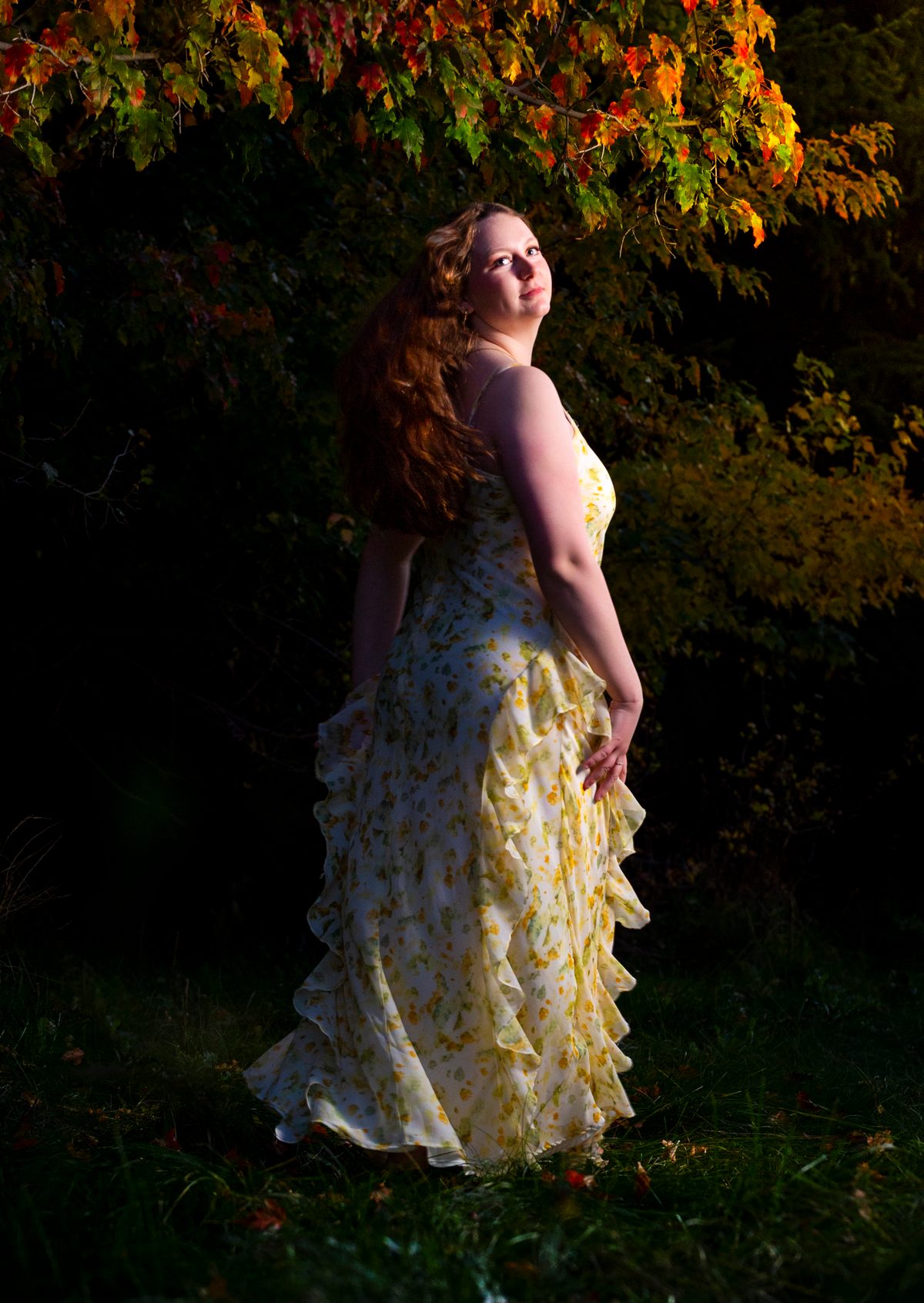 Senior portrait of a young woman in a flowing yellow dress standing among autumn leaves at dusk