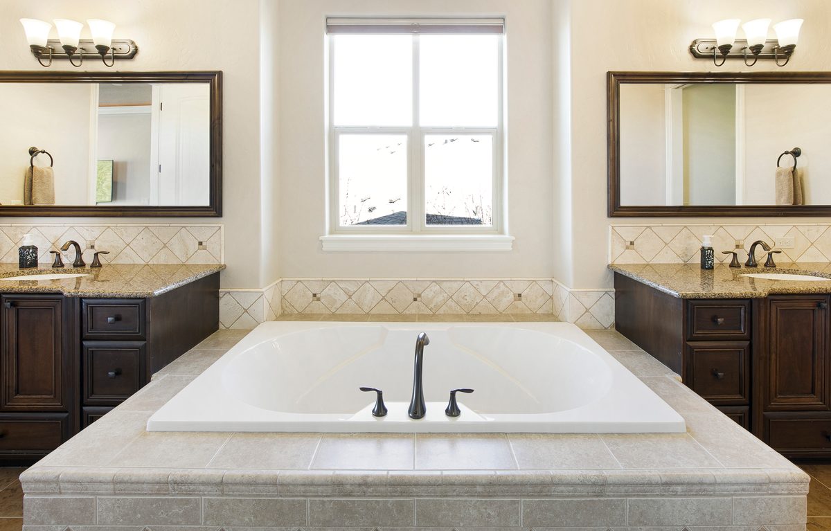 Primary bathroom with a soaking tub under a window between two dark vanities