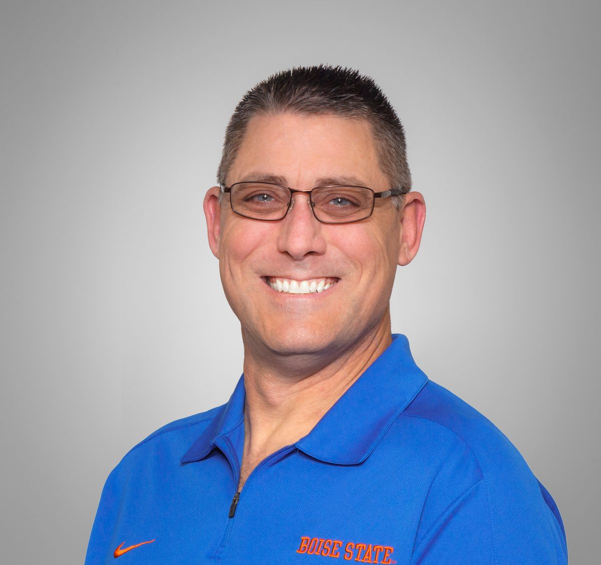 Studio headshot of a smiling man in glasses and a blue polo on a gray background