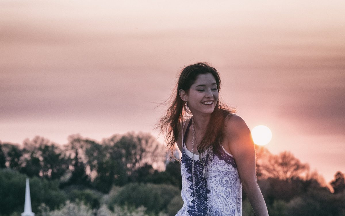 A smiling young woman in a field with the setting sun and a church steeple behind her