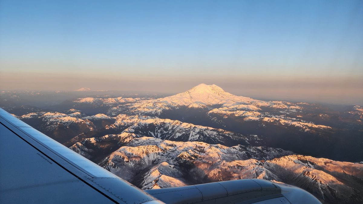 A snow-capped volcano rising above a sea of clouds, seen from an airplane window at sunrise