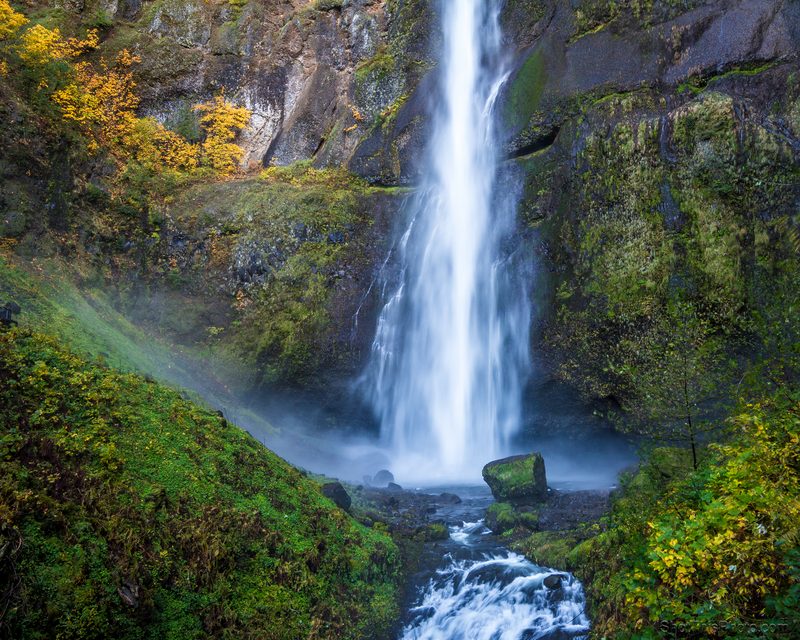 A tall waterfall plunges down a mossy cliff into a misty pool with autumn color above.