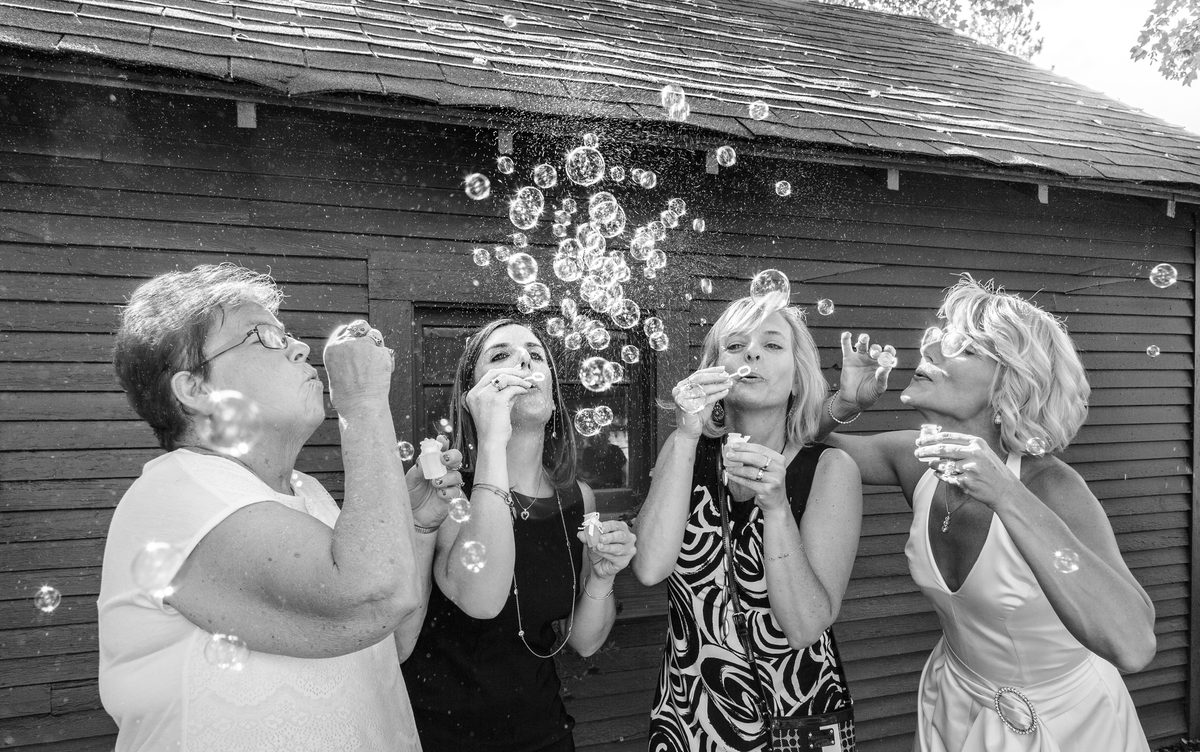 Black-and-white photo of four women blowing bubbles in front of a barn at a wedding