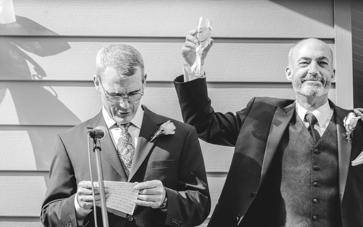 Black-and-white photo of two men in suits giving a wedding toast, one raising his hand