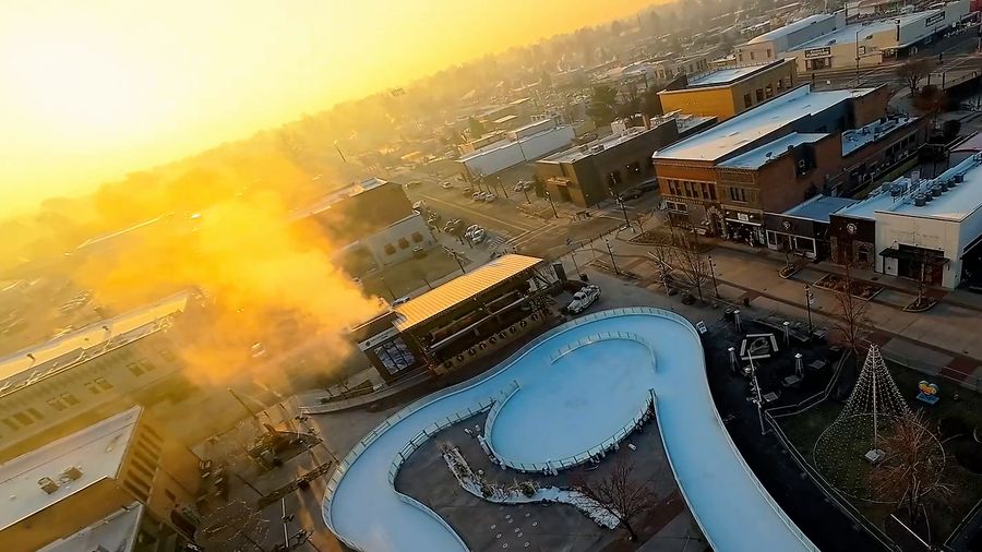 FPV drone circles a downtown ice ribbon as steam drifts through golden sunrise light, then skims a lake