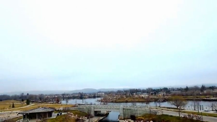 FPV drone glides over a riverside park with colorful poles, then follows a rocky river channel under a bridge