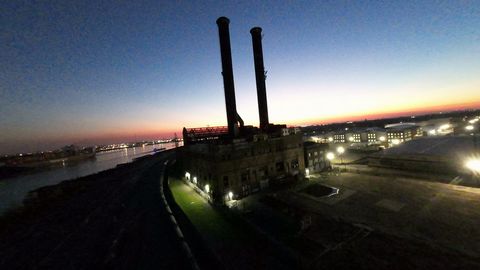 FPV flight around the two tall chimneys of an old riverside power plant at dusk, city lights beyond